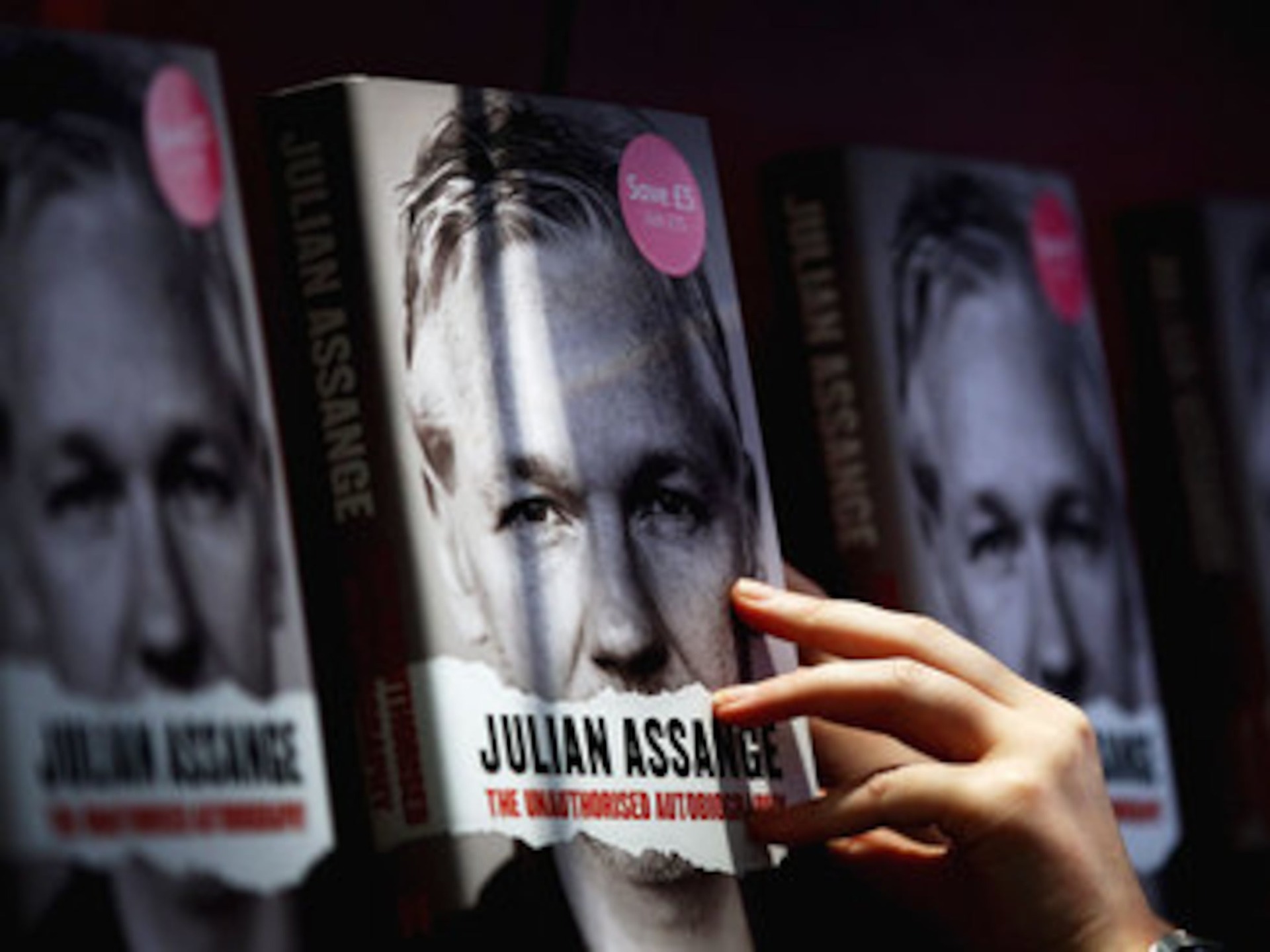 Mano sfiora copertina di libri con volto di Julia Assange, scaffale sfocato sullo sfondo.